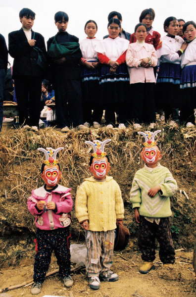 苗族的节庆上，三个小孩带上孙悟空的面具。上方女孩穿着手工染蓝的裙子在旁观礼。贵州六盘水